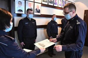 Naczelnik Wydziału Kadr przekazuje rozkaz Komendantowi Wojewódzkiemu, w tle policjanci, którzy oczekują na ich wręczenie.