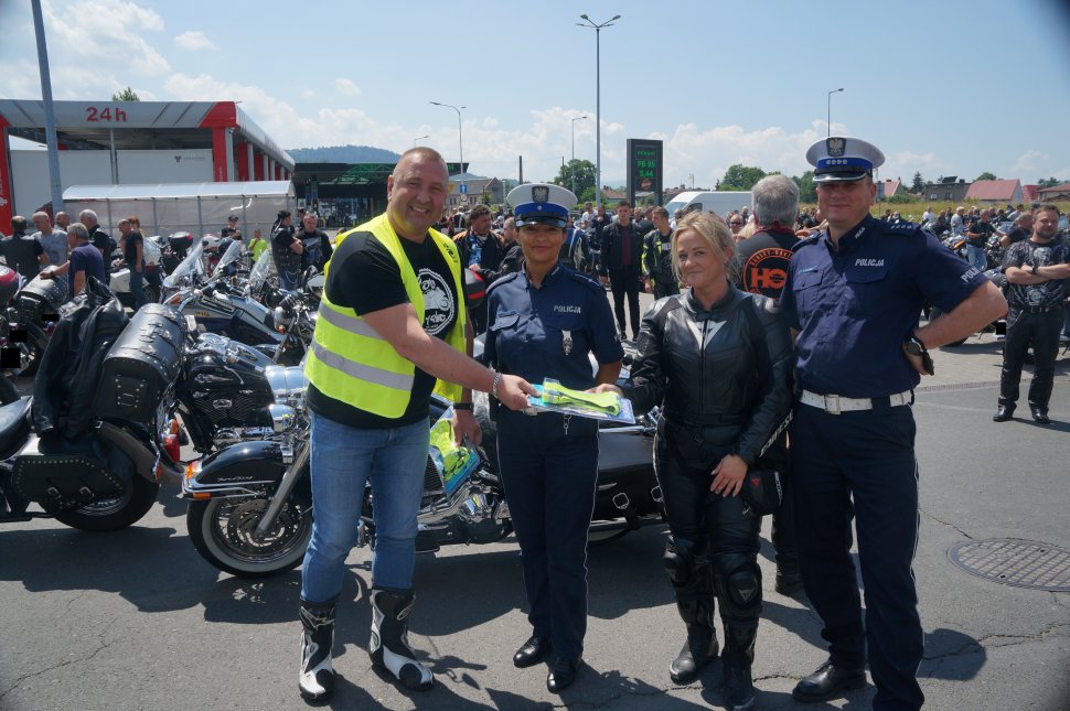 Stoją motocykliści i policjanci obok siebie motocyklista wręcza szelki odblaskowe innemu motocykliści kobiecie w tle motocykle orqaz inni motocykliści