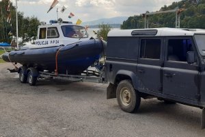 widoczny fragment samochodu z wózkiem do przewożenia łodzi na którym znajduje się policyjna motorówka w tle góry i jezioro