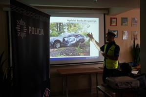 policjant stoi bokiem i ręką pokazuje coś na wyświetlonym slajdzie, slajd pokazuje samochód po wypadku