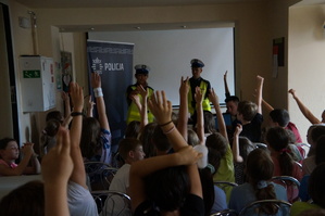 uczestnicy spotkania siedzą tyłem na krzesłach przed nimi stoi policjantka i policjant wielu z dzieci zgłasza się poprzez podniesienie ręki