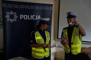 na środku sali stoi policjantka i policjant policjant trzyma w ręce odblaskową zawieszkę w kształcie raqdiowozu