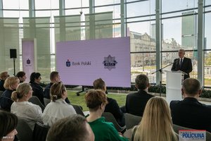 na zdjęciu minister spraw wewnętrznych przy mównicy przed publicznością obok białego dużego ekranu