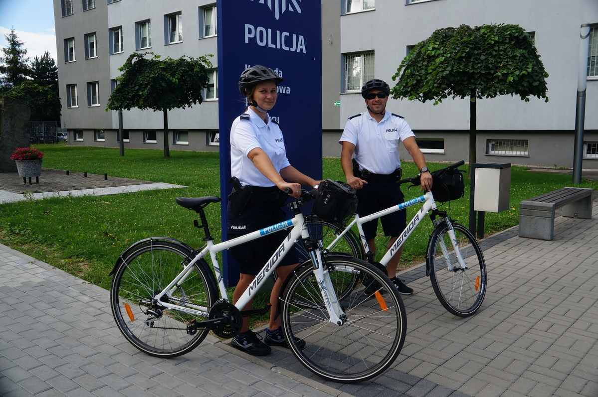 policjantka i policjant z rowerami stoją obok pylonu komendy powiatowej policji w Żywcu w tle budynek komendy