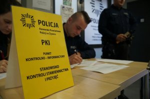 na pierwszym planie żółta karta informacyjna dotycząca stanowiska przyjęć w tle policjanci