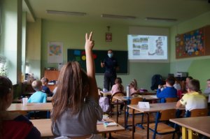 dziewczynka siedzi tyłem w ławce i zgłasza się w tle reszta klasy i policjantka