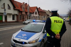 zdjęcie kolorowe policjant stoi tyłem obok radiowóz w tle ulica i skrzyżowanie