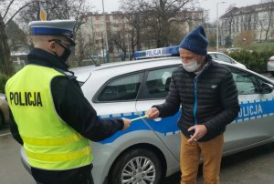 policjant stoi bokiem obok niego meżczyzna policjant daje mu odblask obok fragment radiowozu w tle bloki