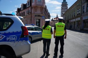 na pierwszym planie stoi policjnat i policjantka tyłem obok zaparkowane radiowozy w tle ulica