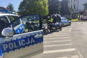 na pierwszym planie fragment radiowozu z otwartymi drzwiami widoczyny napis policja w tle ulica i policjanci na motorach