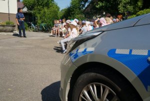 na pierwszym planie fragment radiowozu w tle policjantka w czasie prelekcji i dzieci które siedzą na ławkach w tle budynek szoły