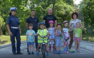 Zdjęcie grupowe obok siebie stoi policjantka dzieci i ich tatusiowie dzieci trzymają hulajnogi, rolki, w tle drzewa, zdjęcie kolorowe