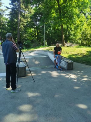 na pierwszym planie stoi mężśczyzna który realizuje spot stoi przed aparatem fotograficznym w tle siedzi męzczyzna i dziecko obok nich stoi hulajnoga w tle drzewa