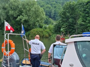 widoczni policjanci na łódce stoją tyłem w tle jezioro brzeg jeziora oraz kajakarze