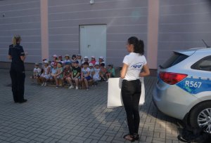 zdjęcie zrobione na placu przedszkola policjantka stoi tyłem przed nią siedzą dzieci obok stoi pracownik z wordu z boku widoczny fragment radiowozu
