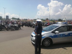 policjantka stoi tyłem obok radiowozu w tle zaparkowane motocykle oraz motocykliści
