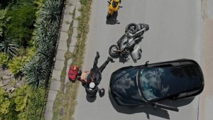 widok z góry na drodze lezy motocyklista obok niego siedzi drugi obok dwa motory  jeden z nich przewrócony z boku stoi samochód