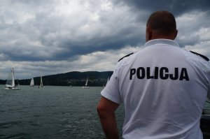 na pierwszym planie policjant stoi tyłem w koszulce z napisem policja w tle jezioro żagłówki i góry