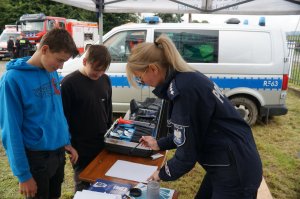 zdjęcie kolorowe zrobione w plenerze stosisko profilaktyczne policyjne, policjantka stoi bokim za stołami przed nią dwóch chłopaków, widoczny policyjny sprzęt i radiowóz