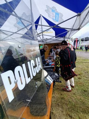 na pierwszym planie policyjna tarcza widoczny napis policja w tle stoisko policyjne przy którym stoją ludzie