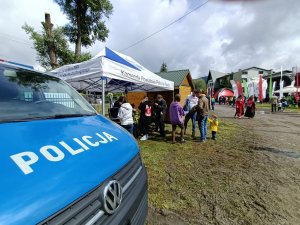 na pierwszym planie widoczny fragment radiowozu widoczny napis policja w tle stosiko policyjne przy którym stoją osoby