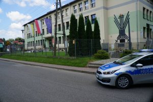 zdjęcie kolorowe po prawej stronie zdjęcia fragment radiowozu resztę zdjęca zajmuje ulica i budynek szkoły