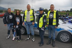 na tle radiowozu stoją motocykliści policjantka i dziecko w tle inni motocykliści i motocykle