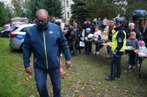 męzczyzna w założonych alkogooglach pokonuje slalom w tle radiowóz policjantka i uczsetnicy imprezy