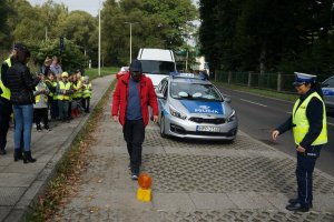 osoba pokonuje slalom w założonych alkogooglach obok stoi policjantka po lewej stronie stoją dzicie w tle radiowóż