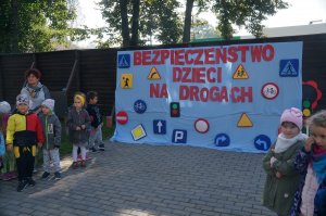 na zewnątrz stoją przedszkolaki na wystawce napis bezpieczeństwo dzieci na drogach