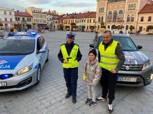 zaparkowany radiowóz oraz samochód radia bielsko obok stoi policjantka redaktor oraz dziecko z założonym odblaskiem