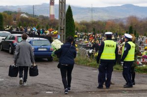 policjanci stoją i rozmawiają z osobą obok widać zaparkowane samochody i liudzi idących na cmentarz w tle cmentarz