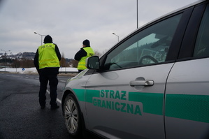 fragment samochodu straży granicznej przed nim tyłem stoi dwóch policjantów