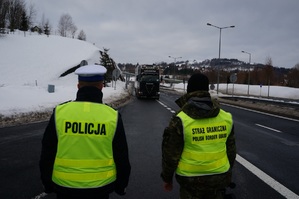 policjant i funkcjonariusz straży granicznej stoją tyłem przed nimi droga po której jedzie tir