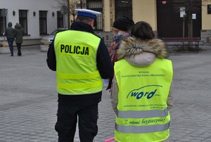 tyłem stoi policjant i pracownik word obok kobieta której wręczają odblask