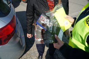 fragment radiowozu policjantki oraz pieszej policjantka trzyma w dłoniach ulotki i odblaskową kamizelkę