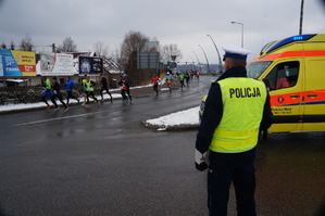 policjant stoi tyłem obok niego ambulans pogotowia w tle biegacze