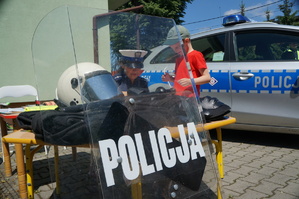 na pierwszym planie policyjna tarcza z napisem policja w tle policjantka pokazuje dziecku jak zabezpiecza się odciski linii papilarnych w tle radiowóz