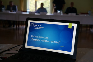 na pierwszym planie pulpit laptopa na którym jest slajd z tytułem debaty bezpieczeństwo w sieci