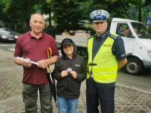 na pierwszym planie stoi mężczyzna obok niego dziecko i policjant mężczyzna i dziecko trzymają odblaski w tle ulica i samochody