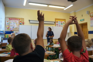 na pierwszym planie zgłaszające się do odpowiedzi dzieci w tle policjantka