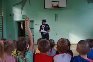 policjantka w czasie prelekcji stoi przodem do dzieci, dzieci siedzą tyłem do obiektywu na ławeczkach zdjęcie kolorowe zrobione w sali gimnastycznej, pozostałe zdjęcia zrobione w sali gimnastycznej