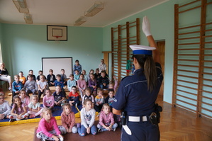 policjantka tyłem prezentuje dzieciom ręczne kierowanie ruchem przez policjanta dzieci siedzą przodem do niej