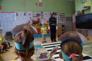 na pierwszym planie siedzą dzieci tyłem mają założone na głowie zrobione policyjne czapki w tle policjantka