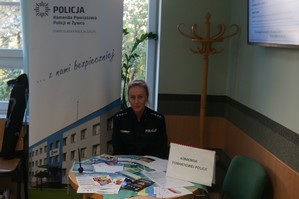 policjantka siedzi przy stole na którym leżą ulotki obok niej baner komendy