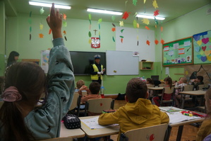 na pierwszym planie uczniowie siedzą tyłem jedna z uczennic zgłasza się przez podniesienie ręki w tle policjantka