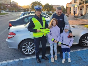 obok siebie stoi policjantka obok niej stoi mężczyzna i dwie dziewczynki wszyscy trzymają odblaski w tle ulica samochody i panorama miasta