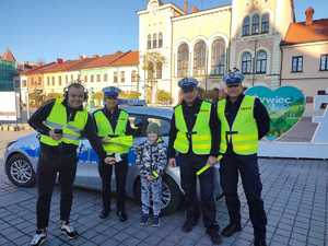 na tle radiowozu stoją policjanci redaktor oraz chłopczyk wszyscy trzymają odblaski w tle rynek miasta