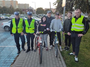 obok siebie stoją policjanci redaktorzy i uczestnicy akcji dwóch z nich ma rowery w tle samochody, ulica i bloki