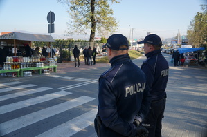 grafika poglądowa policjanci stoją w rejonie przejścia dla pieszych w tle cmentarz i osoby